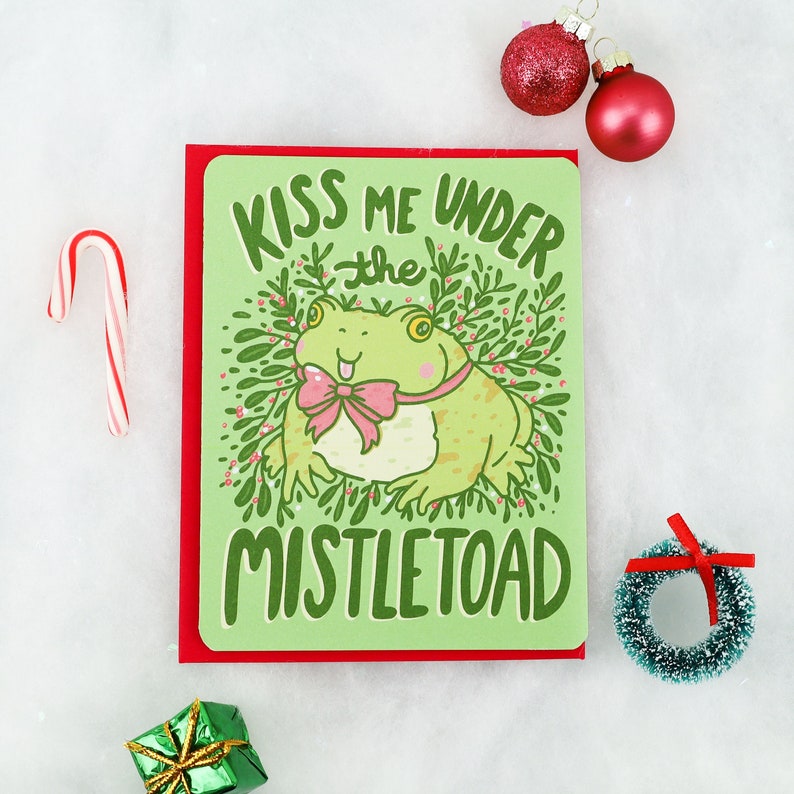 May include: A green greeting card with a red border. The card features a cartoon frog wearing a pink bow and surrounded by green leaves. The text on the card reads "Kiss me under the mistletoe".