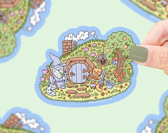 Puede incluir: Una pegatina caprichosa con una ilustración de dibujos animados de un agujero de hobbit con un mago y un gato leyendo un libro. La escena incluye un árbol con una pajarera, flores y una regadera. La pegatina está delineada en azul.