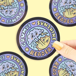 May include: Round sticker with a black border featuring a cartoon illustration. The design includes a cloud with rain and lightning, a sun, and the words "Grumpy Sunshine Reader". The sticker is held by a hand.