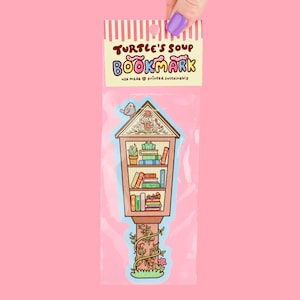 May include: A bookmark featuring a whimsical illustration of a miniature library, complete with shelves of books, plants, and a small bird perched on top. The bookmark is packaged in a clear plastic sleeve with the text "Turtle's Soup Bookmark".