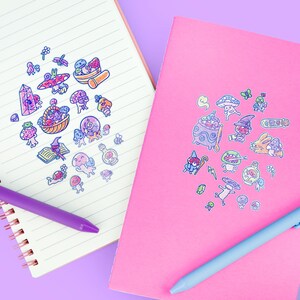 May include: Two notebooks, one lined and one pink, adorned with colorful mushroom and fantasy-themed stickers. A purple pen rests on the lined notebook, and a blue pen on the pink one. The stickers feature whimsical illustrations of mushrooms, potions, and magical creatures.