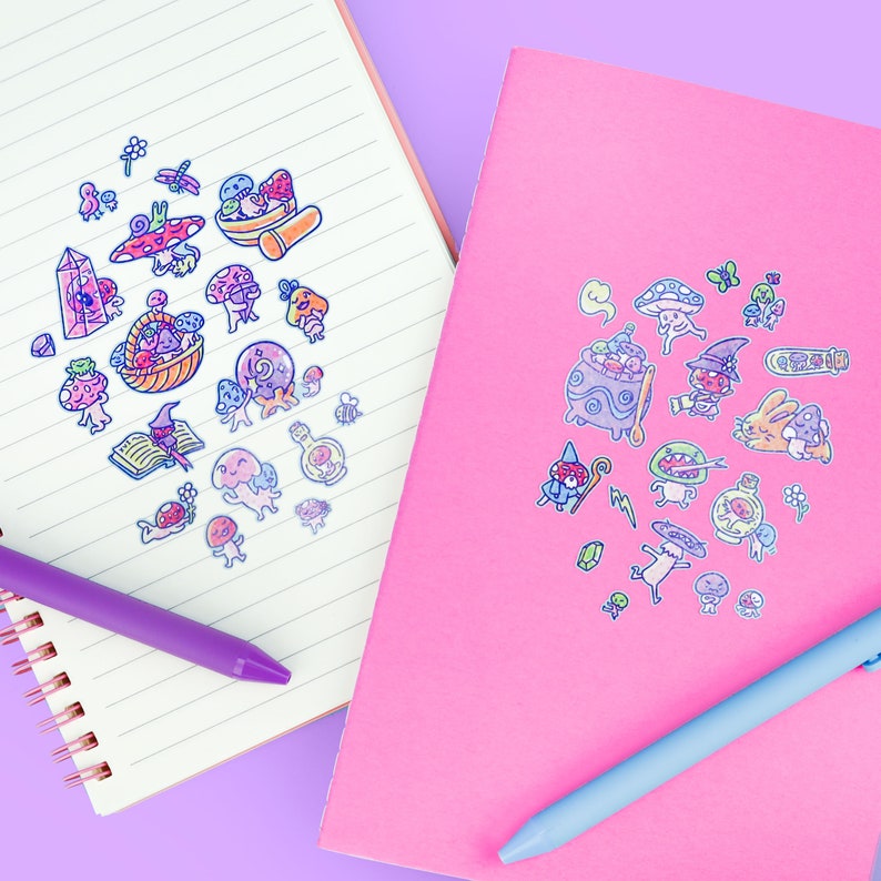 May include: Two notebooks, one lined and one pink, adorned with colorful mushroom and fantasy-themed stickers. A purple pen rests on the lined notebook, and a blue pen on the pink one. The stickers feature whimsical illustrations of mushrooms, potions, and magical creatures.