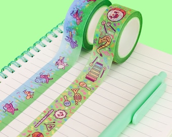 May include: Two rolls of green washi tape with colorful illustrations. One roll features musical instruments and the other features bugs and flowers. The tape is on a white notebook with green spiral binding.