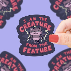 May include: A sticker with a cartoon cat illustration. The cat is gray and has an angry expression, with yellow lightning bolts around it. The sticker reads "I AM THE CREATURE FROM THE FEATURE" in pink and white text. The website www.turtlessoup.com is also visible.
