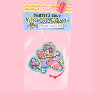 May include: Air freshener in a clear plastic package. The air freshener features a cartoon bear wearing sunglasses and a scarf, riding in a blue, red, and yellow airplane. The packaging reads "Turtle's Soup Air Freshener Fresh Take-Off."
