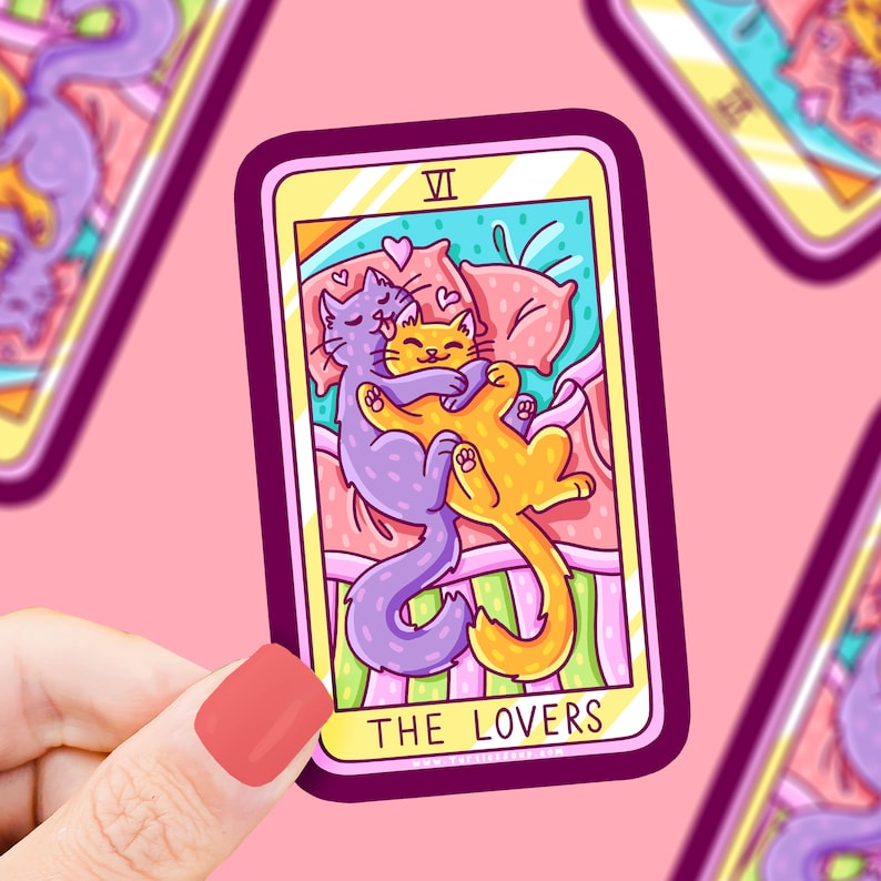 May include: A tarot card with a purple and orange cat couple cuddling on a bed. The card is titled "The Lovers" and has the Roman numeral VI in the top right corner.