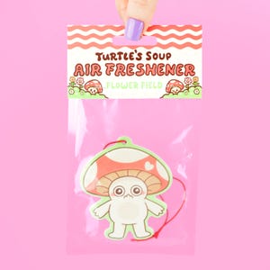 May include: A pink package with a mushroom-shaped air freshener. The air freshener has a red and white mushroom cap with a green outline and a cartoon face. The package reads "Turtle's Soup Air Freshener Flower Field."