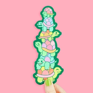 May include: A sticker of a stack of five cartoon turtles, each with a different colored shell. The turtles are stacked on top of each other, with the smallest turtle on top. The turtles are all smiling and have green bodies.