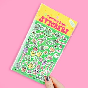 May include: A sheet of colorful stickers featuring gardening themed illustrations. The stickers include a watering can, a gnome, a shovel, a rake, a pair of gardening gloves, a frog, a tomato, a cucumber, a sunflower, a bee, a butterfly, and more. The stickers are packaged in a clear plastic bag with a green and yellow background. The text on the bag reads "Turtle's Soup Stickers".