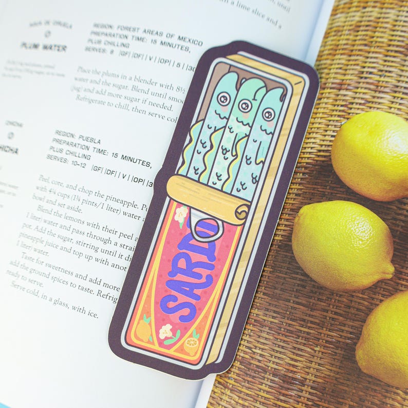 May include: A colorful sticker featuring a cartoon sardine can with three smiling fish. The can is labeled "SARIO" in a stylized font, with floral and citrus accents. The sticker is placed on a cookbook page next to lemons.