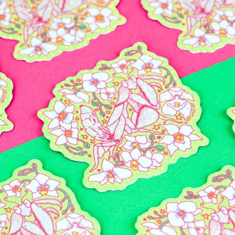 May include: A green sticker with a white praying mantis surrounded by white flowers and yellow stars. The sticker has the text "@tiny.tarus.shop" in the bottom right corner.