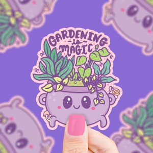 May include: A purple cauldron with a smiling face and a frog peeking out of the top. The cauldron is filled with green plants and the text "GARDENING -is- MAGIC" is written above the cauldron.