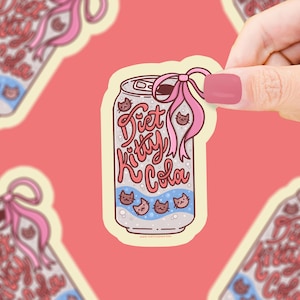 May include: A sticker featuring a cartoon soda can design. The can is labeled "Diet Kitty Cola" in pink and blue lettering, with cat faces and a pink bow. The sticker is held by a hand against a pink background.