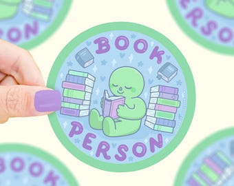 Puede incluir: Adhesivo redondo con un alienígena verde leyendo un libro, rodeado de pilas de libros y las palabras "BOOK PERSON". El diseño presenta un fondo azul claro con estrellas y pequeños libros.