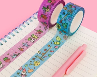 May include: Two rolls of washi tape with a gardening theme. One roll is purple with green and yellow illustrations of plants and vegetables. The other roll is blue with green and yellow illustrations of gardening tools and flowers. The tape is unrolled on a notebook with lined pages.