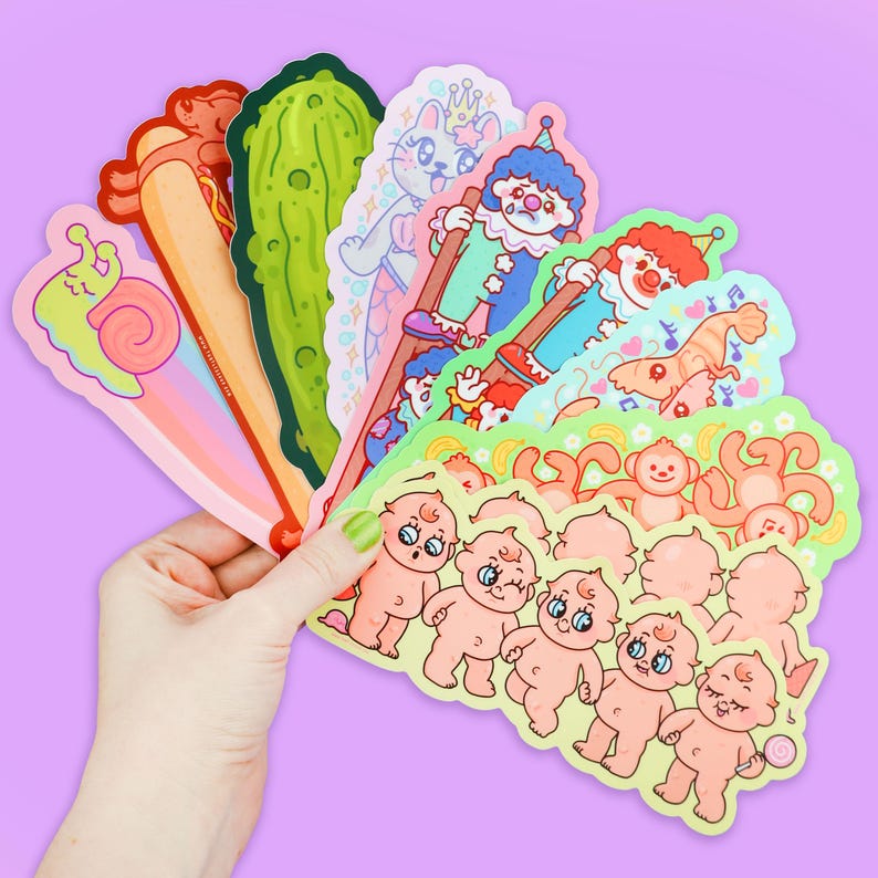 May include: A set of colorful stickers featuring various cartoon characters, including a snail, a pickle, a mermaid, a clown, a monkey, and a baby. The stickers are arranged in a fan shape and are held in a hand.