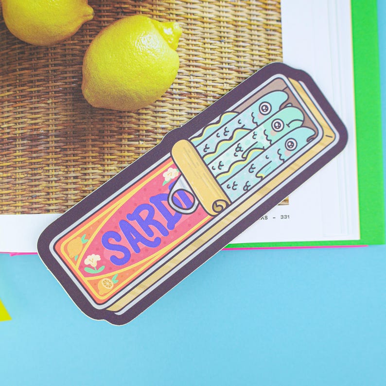 May include: A sticker featuring a sardine can illustration with the word "SARDO" in purple and blue. The can contains three cartoon sardines. Two yellow lemons are in the background, with a woven texture.