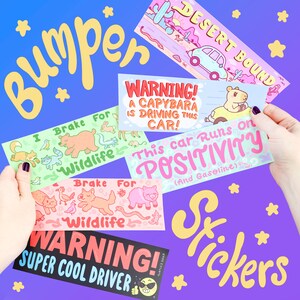 May include: A set of colorful bumper stickers with various designs. The stickers feature illustrations of animals, text, and a cartoon character. The text on the stickers includes "I Brake For Wildlife", "WARNING! A Capybara Is Driving This Car!", "This Car Runs On Positivity (And Gasoline)", and "WARNING! Super Cool Driver".