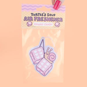 May include: Air freshener in a clear plastic package. The air freshener has a cartoon snail and two pink square candies with heart and star patterns. The package reads "Turtle's Soup Air Freshener Kawaii Candy."