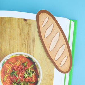 May include: A bread-shaped bookmark with a brown border and white details, placed on a book with a green spine. The book is open to a page with a bowl of soup and a wooden background. The background is blue.