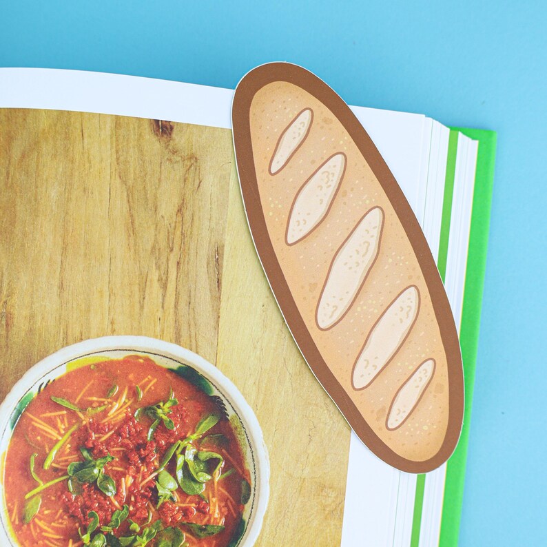 May include: A bread-shaped bookmark with a brown border and white details, placed on a book with a green spine. The book is open to a page with a bowl of soup and a wooden background. The background is blue.