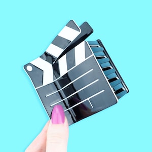 May include: A black and white film clapperboard with a glossy finish. The clapperboard is held against a light blue background. The clapperboard is open, revealing the interior. The clapperboard is a prop used in filmmaking.