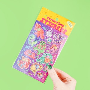May include: A pack of colorful stickers featuring dancing dinosaurs, musical notes, and a disco ball. The stickers are in a clear plastic sleeve with the text "Turtle's Soup Stickers" in pink and yellow. The background is a vibrant green.