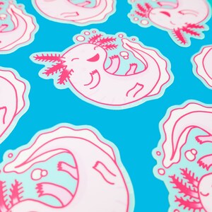 May include: Pink and blue axolotl stickers with a cartoon axolotl design. The axolotl is smiling and has a pink body with blue fins and gills. The stickers are on a blue background.