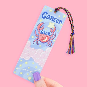 May include: A light blue bookmark featuring a cartoon crab with a star and crescent moon design. The word "Cancer" is written in blue script at the top. The bookmark has a rainbow tassel attached to the top.