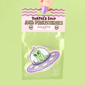 May include: A green alien in a purple spaceship air freshener with the text "Turtle's Soup Air Freshener Galactic scent".