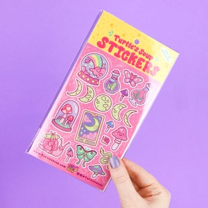 May include: A sheet of pink stickers with a variety of whimsical designs, including moons, mushrooms, a snail, a butterfly, a potion bottle, and a tarot card with the text "The Moon". The stickers are packaged in a clear plastic sleeve with the text "Turtle's Soup Stickers" printed on the front.