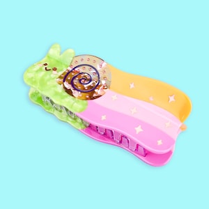 May include: A colorful plastic hair clip shaped like a snail with a green, orange, and pink design. The snail has a sparkly shell with a purple swirl and a cute face.
