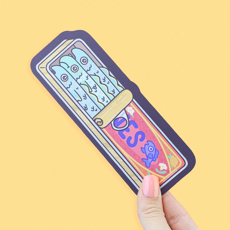 May include: A whimsical sticker featuring a sardine can illustration. The can is red with blue text and a fish graphic. Inside are three cartoon fish with blue bodies and large eyes. The sticker is held against a yellow background.