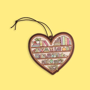 May include: Heart-shaped wooden ornament featuring a bookshelf design. The shelves are filled with colorful books, plants, and decorative items. A brown string is attached for hanging. The ornament is set against a yellow background.