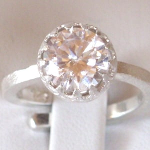 Könnte beinhalten: Ein silberner Ring mit einem großen, klaren, runden Edelstein, der in einem zarten, gewellten Heiligenschein gefasst ist.