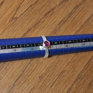 Könnte beinhalten: Ein silberner Ring mit einem kleinen, runden, dunkelrosa Edelstein, der in der Mitte eingesetzt ist. Der Ring wird auf einem Ringmaßstab gemessen und zeigt eine Größe 7.