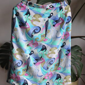May include: A skirt featuring an abstract pattern with swirling shapes in shades of turquoise, blue, pink, and olive green on a white background. The garment has an elasticized waistband and a straight silhouette, hanging from a wooden hanger.