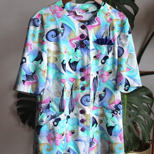 May include: A short-sleeved button-down shirt featuring an abstract pattern in shades of blue, pink, green, and tan. The shirt has a round neckline, five dark purple buttons down the front, and two lower pockets along with a small chest pocket.
