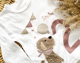 May include: White long-sleeved shirt with a brown dog, pink number two, and the name "Sophie" in pink lettering. The shirt has a pink and beige bunting and a pink heart.