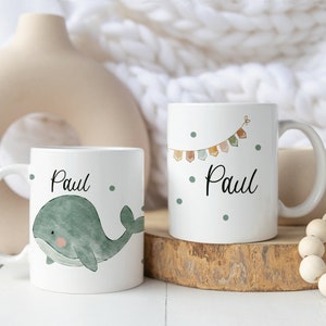 May include: Two white ceramic mugs with the name "Paul" on them. One mug features a green whale illustration, while the other has a colorful bunting design. Both mugs have a white background with small green dots.
