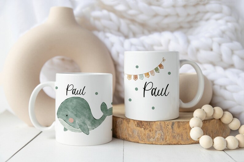 May include: Two white ceramic mugs with the name "Paul" on them. One mug features a green whale illustration, while the other has a colorful bunting design. Both mugs have a white background with small green dots.