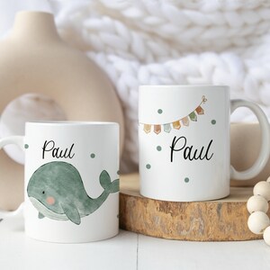 May include: Two white ceramic mugs with the name "Paul" printed on them. One mug features a green whale illustration, while the other has a colorful bunting design.