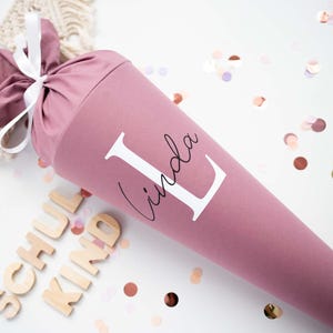 Könnte beinhalten: Eine malvenfarbene Schultüte mit dem Namen "Linda" in Weiß aufgedruckt. Die Tüte ist mit einem weißen Band gebunden. Holzbuchstaben schreiben "SCHUL KIND" neben der Tüte. Konfetti in verschiedenen Farben sind verstreut.