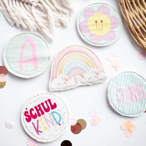 Könnte beinhalten: Eine Sammlung farbenfroher, gestickter Aufnäher mit verschiedenen Designs. Dazu gehören ein Regenbogen, eine Blume mit lächelndem Gesicht und Aufnäher mit dem Buchstaben "A" und dem Namen "ANNI". Konfetti und ein Weidenkorb sind ebenfalls zu sehen.