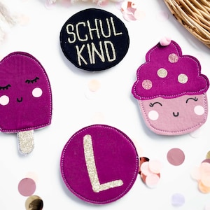 Könnte beinhalten: Eine Sammlung von verspielten Filzaufnähern in Rosa- und Lilatönen. Die Aufnäher zeigen ein Eis am Stiel, einen Cupcake, einen Kreis mit dem Buchstaben "L" und einen marineblauen Kreis mit dem Text "SCHUL KIND". Dekorative Konfetti umgeben die Aufnäher.