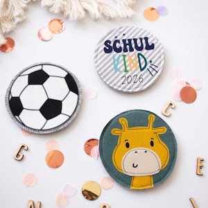 Könnte beinhalten: Drei runde Stoffaufnäher mit verschiedenen Motiven: ein schwarz-weißes Fußballmuster, ein grau-weiß gestreifter Hintergrund mit dem Text SCHUL KIND 2026 und eine gelbe Giraffenillustration auf einem dunkel-petrolfarbenen Hintergrund.