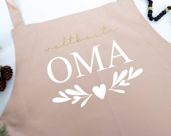 Könnte beinhalten: Eine hellrosa Schürze mit dem Text "weltbeste OMA" in goldener Schrift und einem weißen Herz und floralem Design.