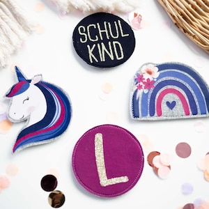 Könnte beinhalten: Eine Sammlung bunter Filzaufnäher, darunter ein Einhorn, ein Regenbogen und ein Kreis mit dem Buchstaben "L". Ein Aufnäher trägt die Aufschrift "SCHUL KIND". Die Aufnäher sind mit Blumen- und Herzdetails verziert, mit Konfetti.