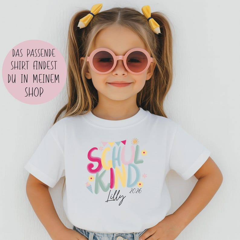 May include: A white t-shirt with the words "School Kind" in colorful letters, with the name "Lilly" and the year "2026" printed below. The shirt is worn by a young girl wearing pink sunglasses.