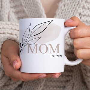 K&ouml;nnte beinhalten: Wei&szlig;e Keramiktasse mit einem schwarzen und braunen Blumenmuster und dem Wort "MOM" in brauner Schrift. Der Text "EST. 2021" befindet sich unter dem Wort "MOM".
